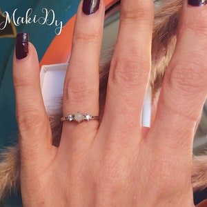 May include: A silver ring with a central, light-colored gemstone flanked by two smaller clear stones on each side. The ring is on a person's finger, with the word "MakiDy" in the upper left corner. The nails are painted a dark burgundy color.