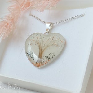 May include: Heart-shaped pendant necklace with a silver chain. The pendant is clear with a tree design and the names "Lara" and "Nick" in black script. The pendant is in a white box with the word "Makidy" in script.