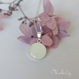May include: A round, off-white pendant necklace with a silver chain. The pendant is set in a silver bezel and hangs against a backdrop of soft pink flowers. The word "Makidy" is written in cursive at the bottom.