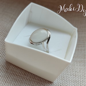 May include: A silver ring with a large, oval, off-white stone. The ring is set in a simple, modern design and is presented in a white box. The ring's band has a split design on either side of the stone.