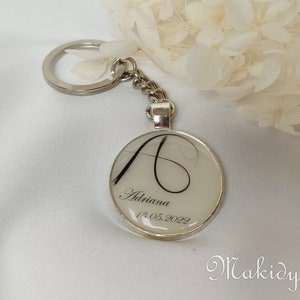 May include: Silver-toned keychain with a round pendant. The pendant features a white background with a black initial "A", the name "Adriana", and the date "13.05.2022". The keychain is attached to a chain and ring.