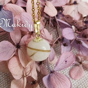 May include: A gold-toned necklace with a round, off-white pendant. The pendant has a gold band and strands of hair. The necklace is set against a backdrop of pink and purple flowers. The word "Makidy" is in the upper left corner.
