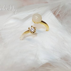 May include: A gold-colored ring with a white gemstone and a small, clear, round stone. The ring has a split shank design. The background is white and fluffy, with the text "Maki Dy" in cursive.