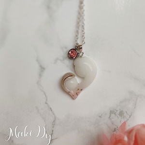 May include: A silver necklace with a pendant featuring a mother and child design. The pendant is white with a pink accent and a small pink gemstone. The necklace is on a white marble surface.