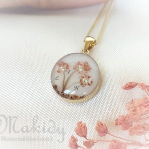 May include: A gold-colored pendant necklace with a round, clear resin charm. The charm contains dried flowers and small crystals, with the letters "L", "I", and "N" visible. The necklace is displayed on a white, textured surface.