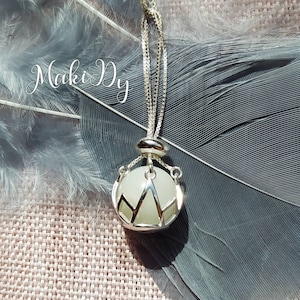 May include: A silver necklace with a round, glowing pendant. The pendant is encased in a silver cage with a mountain-like design. The necklace is displayed on a gray feather and a pink fabric. The text "Maki Dy" is in the upper left corner.