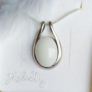 May include: A silver pendant necklace featuring a teardrop-shaped frame holding a white, oval-shaped stone. The necklace is displayed on a white surface. The brand name "Maki Dy" is written in cursive at the bottom.