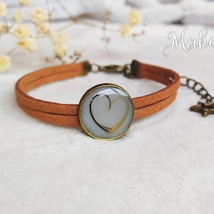 May include: A tan suede bracelet with a round, bronze-colored charm featuring a heart design. The bracelet has a double-strap design and an adjustable chain. The brand name "MakiDy" is written in cursive on the upper right.