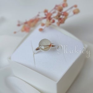 May include: A silver ring with a clear, round, resin-filled stone containing a small, dried plant element. The ring is set on a white surface, with the word "Makidy" in cursive script. A blurred floral arrangement is in the background.