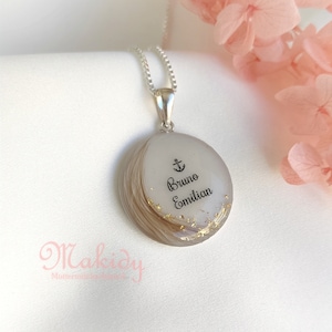May include: A silver pendant necklace with a circular, layered design. The pendant features the names "Bruno" and "Emilian" with an anchor symbol, and gold flakes. The necklace is on a white background.