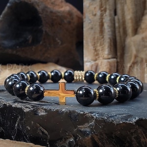 May include: Black beaded bracelet with a gold cross charm. The bracelet is made with black onyx beads and gold spacers.