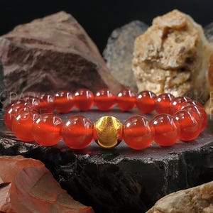 May include: A red carnelian gemstone bracelet with a gold bead accent. The bracelet is made of round beads and is shown on a dark background.