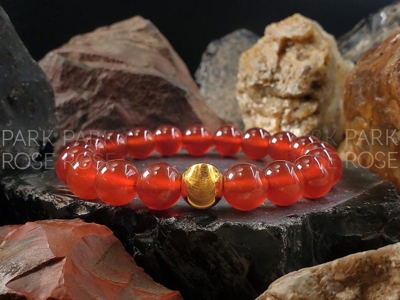 May include: A red carnelian gemstone bracelet with a gold bead accent. The bracelet is made of round beads and is shown on a dark background.