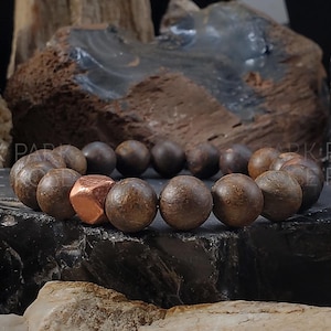 Könnte beinhalten: Ein braunes Holzperlenarmband mit einem kupferfarbenen Metallanhänger. Das Armband liegt auf einer dunkelgrauen Oberfläche.
