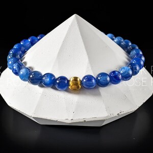 May include: A blue gemstone bracelet with a gold bead accent. The bracelet is made of round beads and is displayed on a white pedestal.