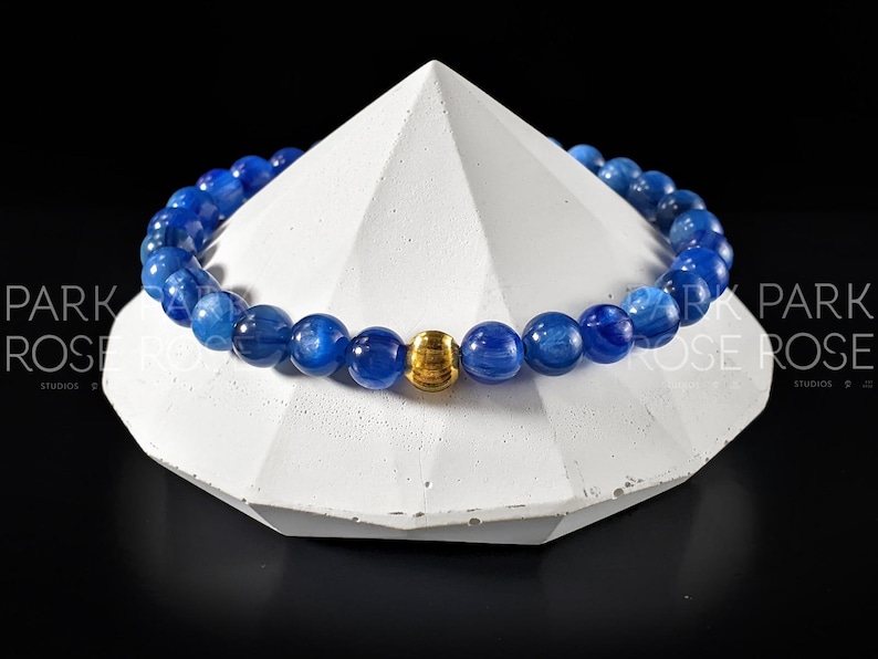 May include: A blue gemstone bracelet with a gold bead accent. The bracelet is made of round beads and is displayed on a white pedestal.
