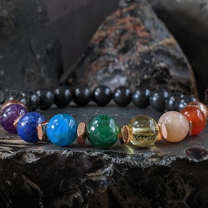 May include: A beaded bracelet featuring a variety of colorful gemstones, including purple, blue, green, yellow, peach, and red. The bracelet also includes black beads and copper-colored spacers. The bracelet is displayed on a dark, textured surface.