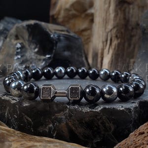May include: A black hematite beaded bracelet with a silver dumbbell charm. The bracelet is on a dark gray background.