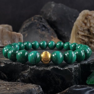 May include: A green malachite gemstone bracelet with a gold bead accent. The bracelet is made of round beads and is strung on a stretchy cord.