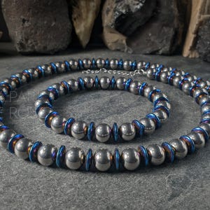 May include: A silver-colored necklace and bracelet set. The jewelry features large, round beads with blue and iridescent accents. The necklace is looped, and the bracelet is a smaller, matching piece. The set is displayed on a gray surface.