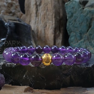 May include: A purple amethyst gemstone bracelet with a gold bead accent. The bracelet is made of round beads and is photographed against a dark background.