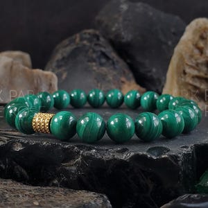 Könnte beinhalten: Ein grünes Malachit-Armband mit goldener Verzierung. Das Armband besteht aus runden Perlen und wird auf einem dunklen Hintergrund gezeigt.