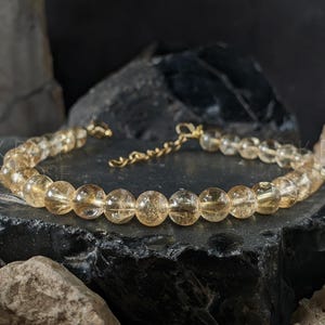 Op de afbeelding: Een armband met ronde, goudgele citrien kralen. De kralen zijn geregen op een gouden ketting met een bijpassende sluiting. De citrien kralen zijn doorschijnend en hebben een warme kleur. De armband ligt op een donkere, getextureerde ondergrond.