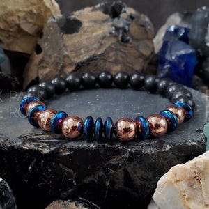 May include: A black and copper bracelet with blue accents. The bracelet is made of round beads and is shown on a dark background.