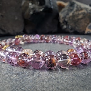 May include: A bracelet made of translucent, round beads in shades of purple, red, and clear. The bracelet is strung on a gold-coloured chain. The beads have inclusions and colour variations. The bracelet is displayed on a grey surface.