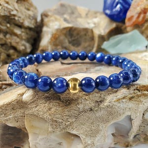 May include: A blue gemstone bracelet with a gold bead accent. The bracelet is made of round beads and is strung on a stretchy cord.