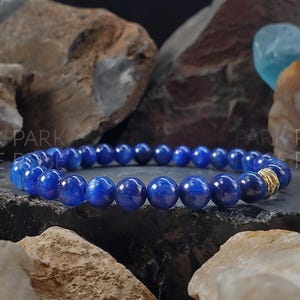 Könnte beinhalten: Ein blaues Edelstein-Armband mit goldenen Akzenten. Das Armband besteht aus runden Perlen und liegt auf einer dunkelgrauen Oberfläche.