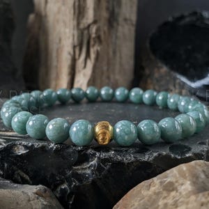 May include: A beaded bracelet featuring round, pale green beads and a gold-colored accent bead. The bracelet is displayed on a dark, textured surface with a natural, earthy background.