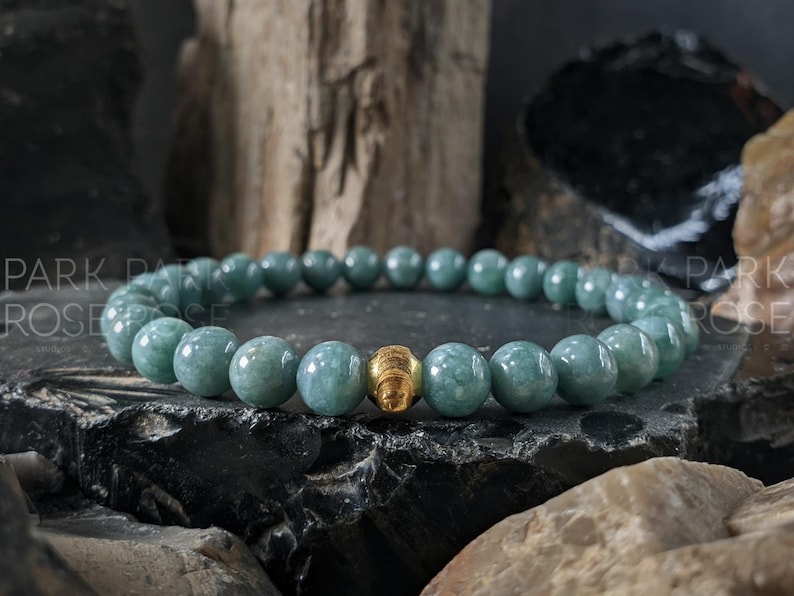 May include: A beaded bracelet featuring round, pale green beads and a gold-colored accent bead. The bracelet is displayed on a dark, textured surface with a natural, earthy background.
