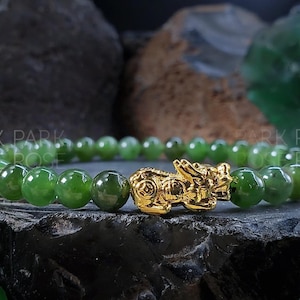 May include: A green jade stone bracelet with a gold charm in the shape of a mythical creature. The bracelet is made of round beads and is approximately 7 inches in length.