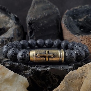 Puede incluir: Un brazalete de piedra de lava negra con un dije de bala dorado con un diseño de cruz. El brazalete está fotografiado sobre un fondo oscuro con rocas.