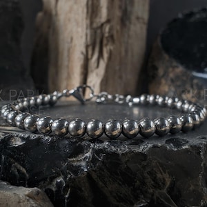 May include: A silver beaded bracelet with a lobster clasp. The bracelet features numerous small, round, metallic beads. The bracelet is displayed on a dark, textured surface. The words "PARK PARK ROSE" are visible in the background.