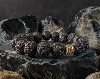 Könnte beinhalten: Ein dunkelbraunes Perlenarmband mit einem goldfarbenen Abstandshalter. Die Perlen haben ein strukturiertes, organisches Aussehen. Das Armband wird auf einer dunklen, strukturierten Oberfläche mit dunklen Felsen im Hintergrund präsentiert.