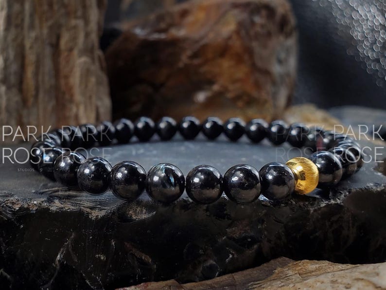May include: Black beaded bracelet with a gold bead accent. The bracelet is made of black gemstone beads and a single gold bead.
