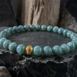 May include: A beaded bracelet featuring round, light teal-colored beads. A single gold-colored bead is centered in the bracelet. The bracelet is displayed on a dark, textured surface.