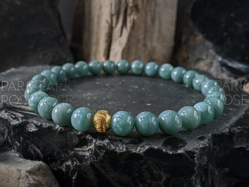 May include: A beaded bracelet featuring round, light teal-colored beads. A single gold-colored bead is centered in the bracelet. The bracelet is displayed on a dark, textured surface.