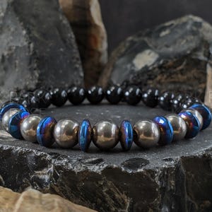 May include: A bracelet with black, silver, and iridescent blue beads. The beads are arranged in a circular pattern. The bracelet is displayed on a dark, textured surface. The "PARK ROSE" logo is visible in the background.