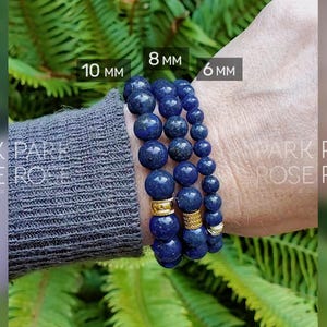 Op de afbeelding: Drie blauwe kralenarmbanden met kralen van 10 mm, 8 mm en 6 mm. De armbanden zitten om een pols, met goudkleurige afstandhouders. De achtergrond is een groene, bladrijke plant.