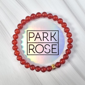 May include: Red carnelian gemstone bracelet with a gold clasp. The bracelet is on a white marble surface with the text "PARK ROSE" in the background.