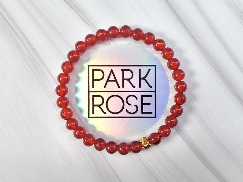 May include: Red carnelian gemstone bracelet with a gold clasp. The bracelet is on a white marble surface with the text "PARK ROSE" in the background.