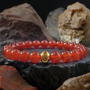 May include: A red carnelian gemstone bracelet with a gold bead accent. The bracelet is made of round beads and is shown on a dark background.