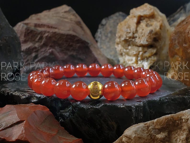 May include: A red carnelian gemstone bracelet with a gold bead accent. The bracelet is made of round beads and is shown on a dark background.