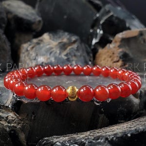 May include: A red carnelian gemstone bracelet with a gold bead accent. The bracelet is made of small, round beads and is shown on a dark background.