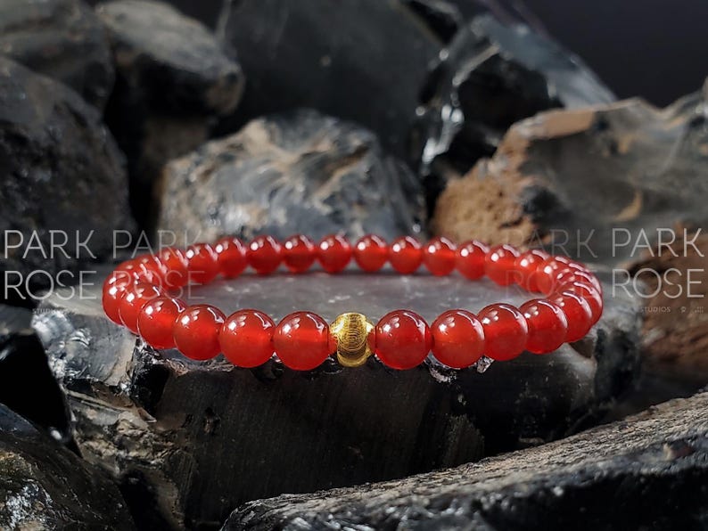 May include: A red carnelian gemstone bracelet with a gold bead accent. The bracelet is made of small, round beads and is shown on a dark background.