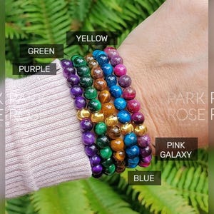 May include: A stack of six colorful beaded bracelets, including purple, green, blue, yellow, and tiger's eye. The bracelets are stacked on a person's wrist. The text "PINK GALAXY" and "BLUE" are visible on the image.