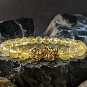 Könnte beinhalten: Armband mit goldgelben Citrin-Edelsteinperlen und einem goldfarbenen Pixiu-Anhänger, einem mythischen chinesischen Wesen. Die Perlen, etwa 1 cm im Durchmesser, liegen auf einer dunklen, strukturierten Oberfläche.